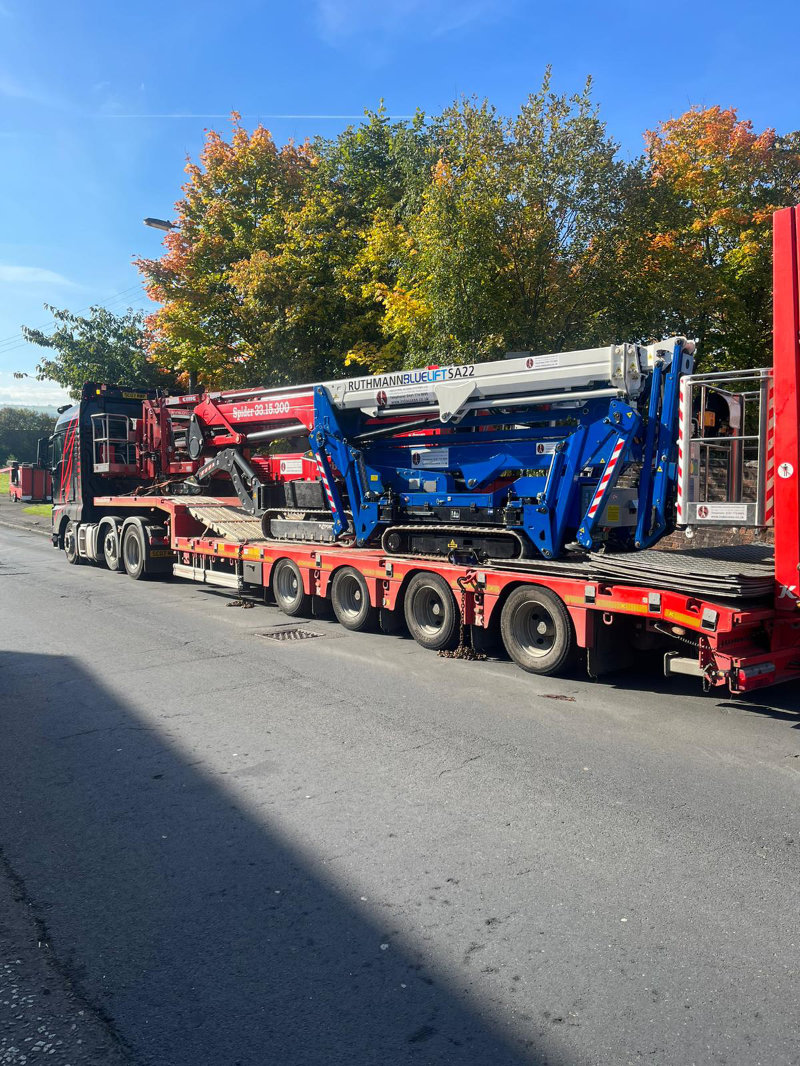 Blue tracked lift being transported on red flatbed trailer truck with autumn trees in background Blue tracked lift being transported on red flatbed trailer truck with autumn trees in background