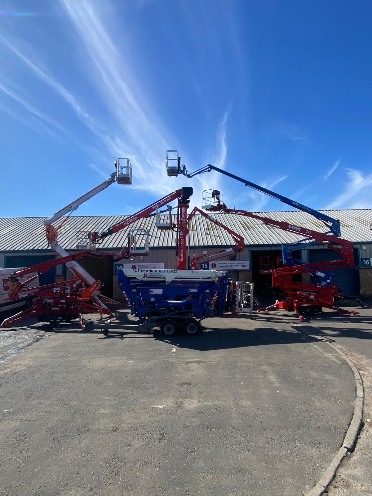 Multiple aerial work platforms and cherry pickers displayed in equipment yard under blue sky with wispy clouds Multiple aerial work platforms and cherry pickers displayed in equipment yard under blue sky with wispy clouds