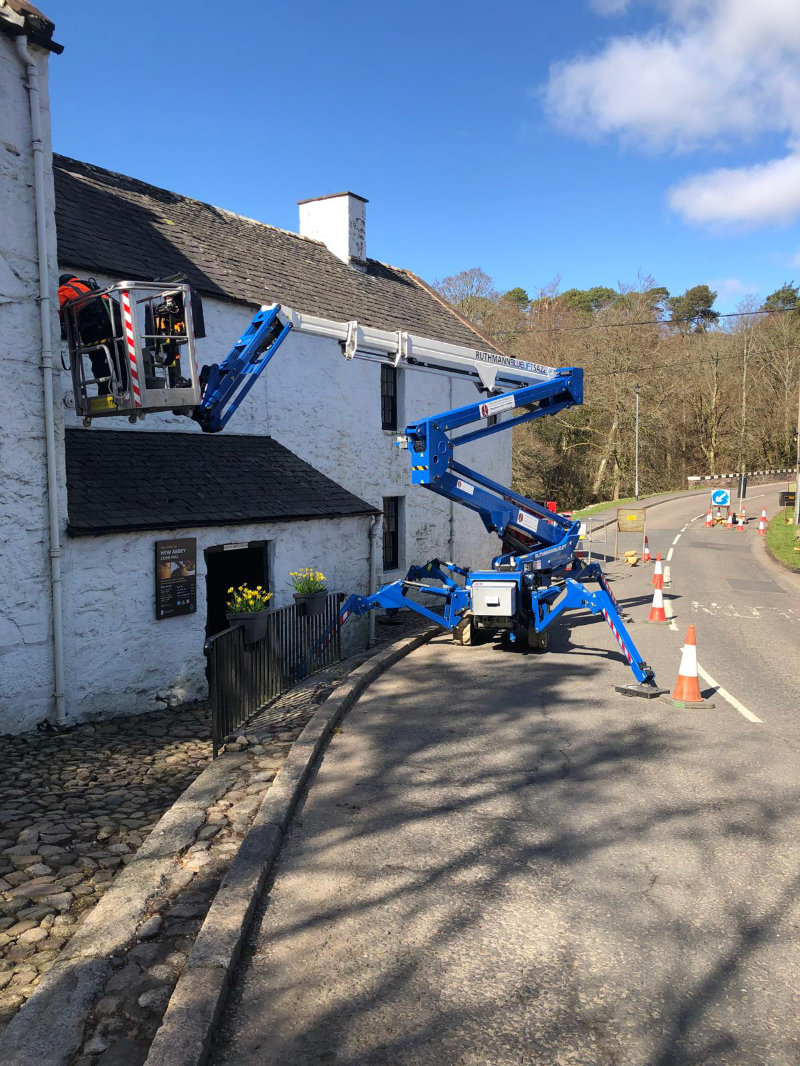 Blue tracked spider lift positioned on road for maintenance work on white cottage with slate roof Blue tracked spider lift positioned on road for maintenance work on white cottage with slate roof