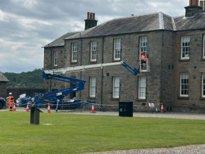 Blue cherry picker extending toward gray stone Georgian building with workers and traffic cones on surrounding lawn
