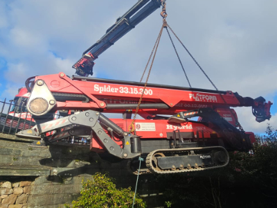 Red tracked spider crane lifting materials with cables against blue sky and stone wall